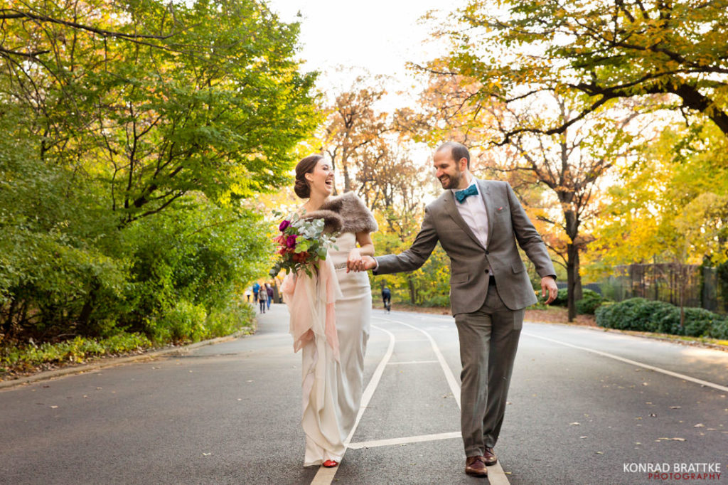 A newlywed couple, dressed in wedding attire, joyfully strolling through Prospect Park before their ceremony.