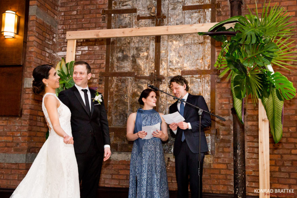 A couple, dressed as bride and groom, exchange vows in front of a brick wall at the 26 Bridge venue.