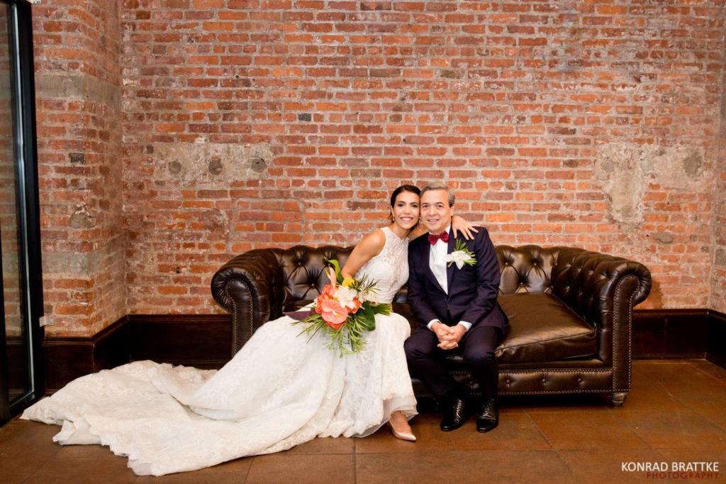 A bride and her dad sitting on a couch in front of a 26 Bridge wall.