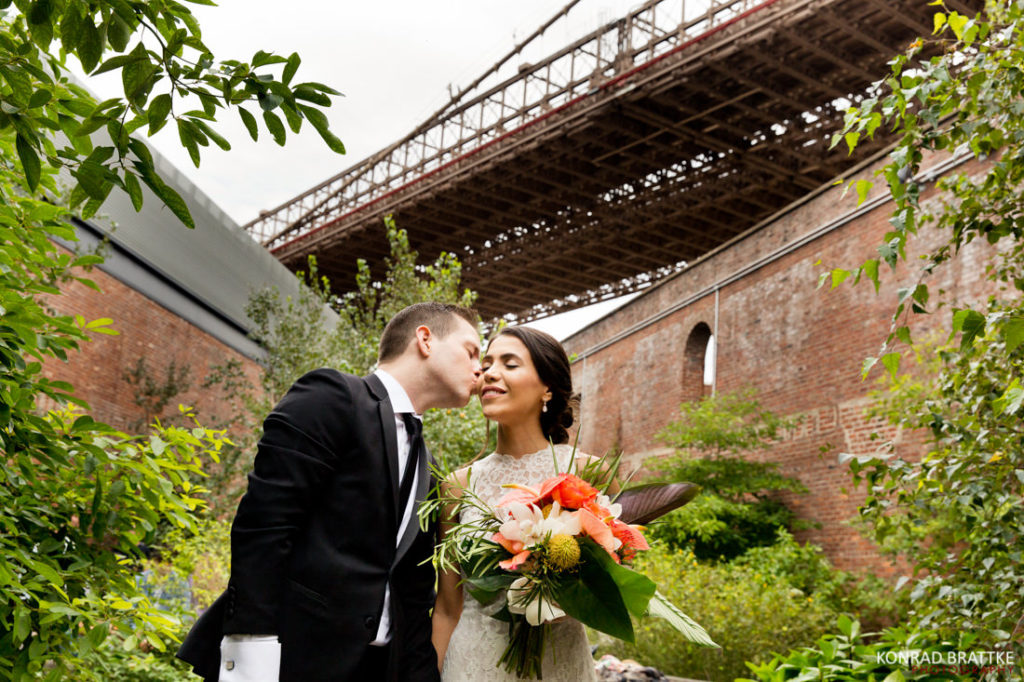 In front of the Brooklyn Bridge, a bride and groom share a tender kiss.