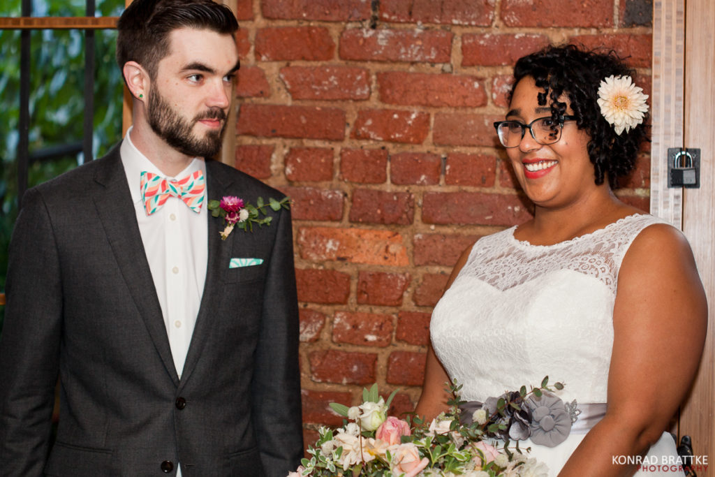 A happy bride and groom smiling in front of a brick wall at their Brooklyn wedding.