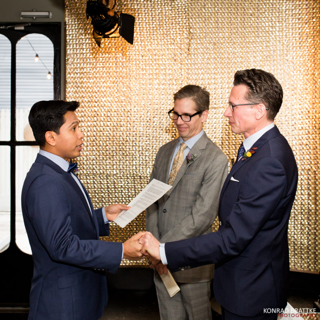Two grooms at Le Fanfare holding hands during their wedding ceremony.