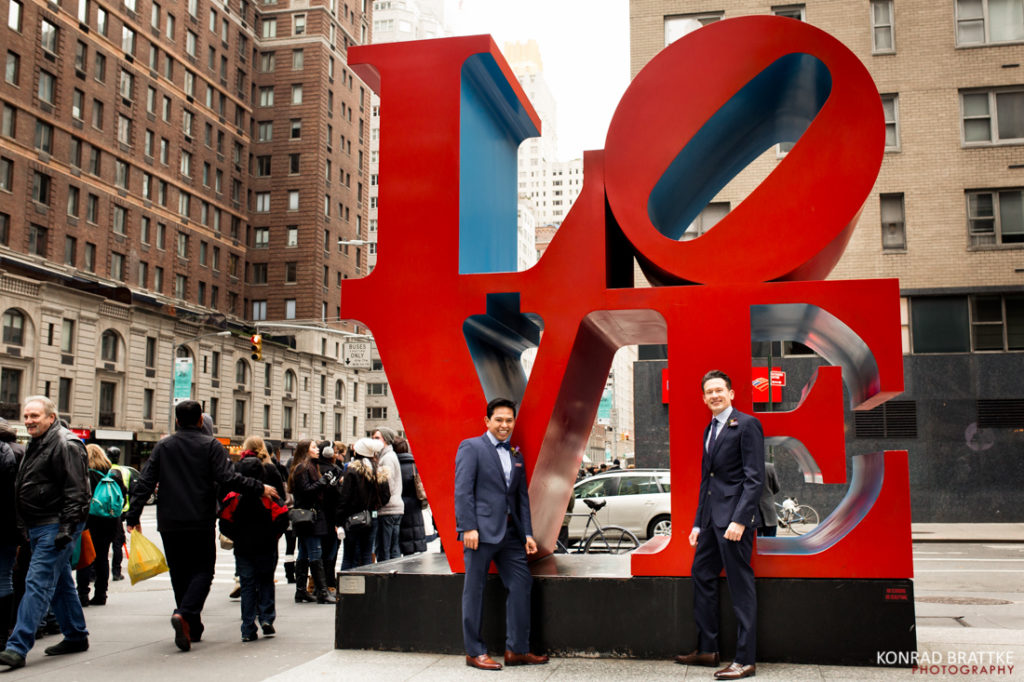 Two grooms standing in front of a large love sculpture