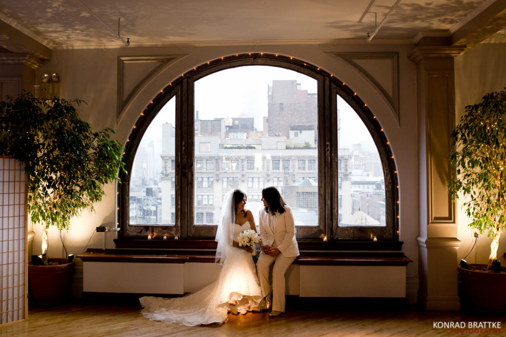 Two brides standing in front of a large window at their Manhattan Penthouse wedding.