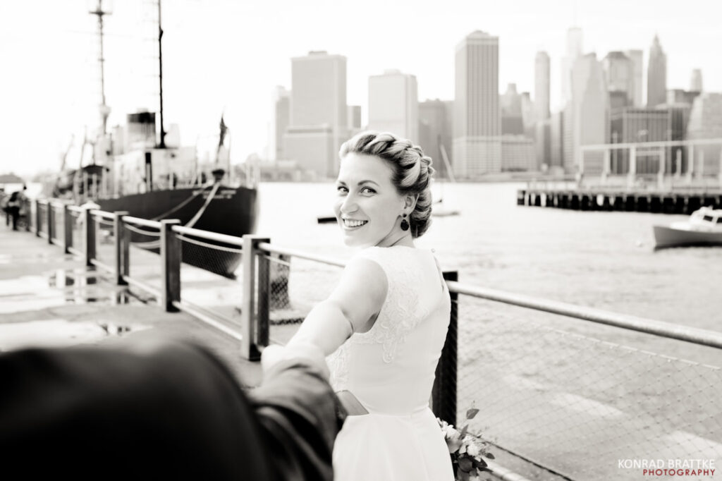 A bride and groom walking on a dock in front of a city skyline.
