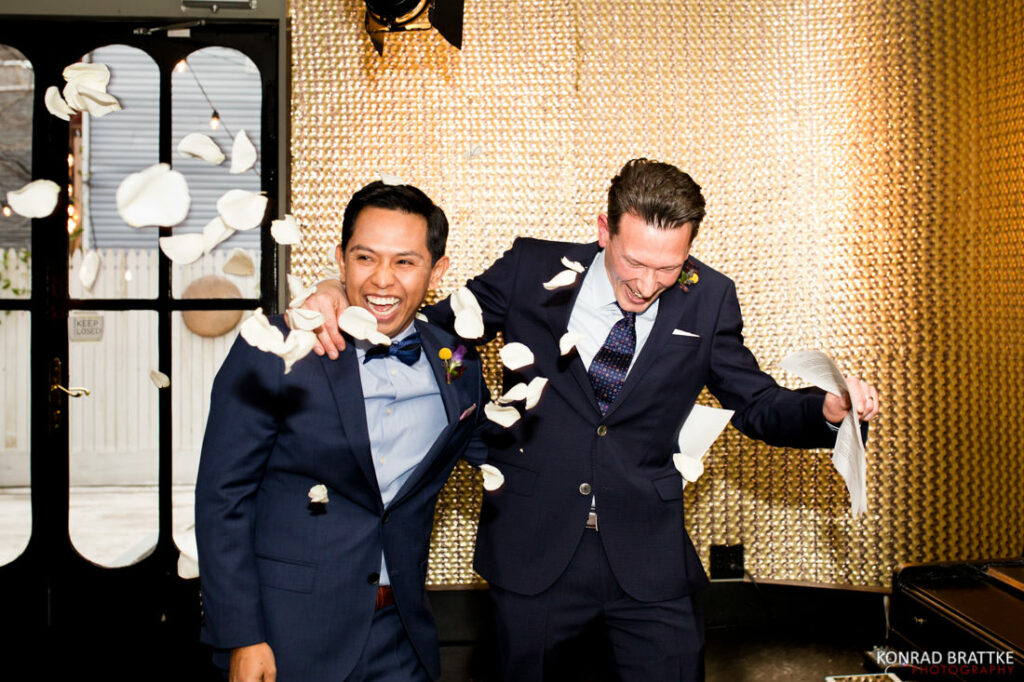 Two grooms in suits are throwing petals at each other during their ceremony.