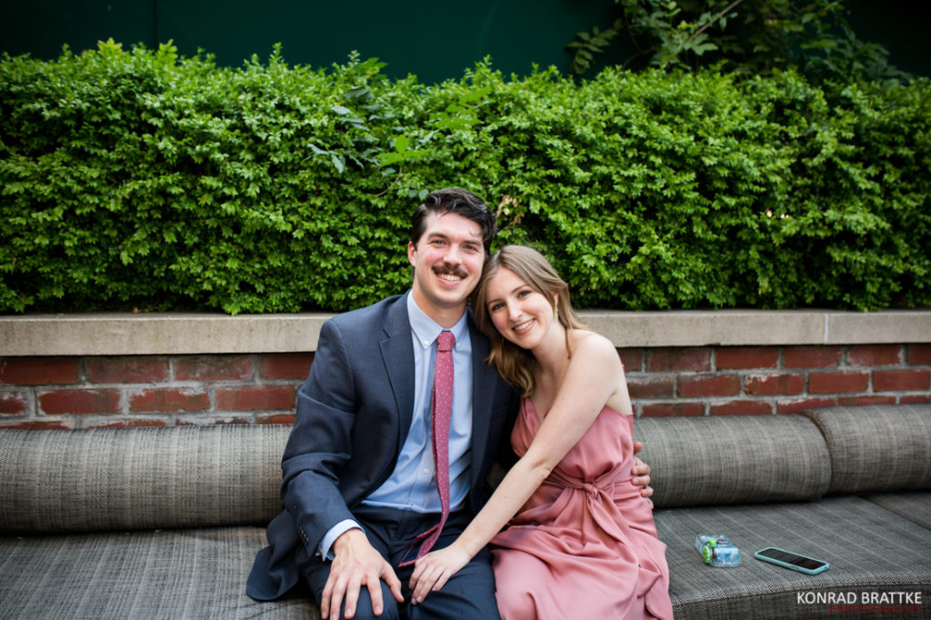 A man and woman sitting on a couch in front of bushes at a Bowery Hotel.