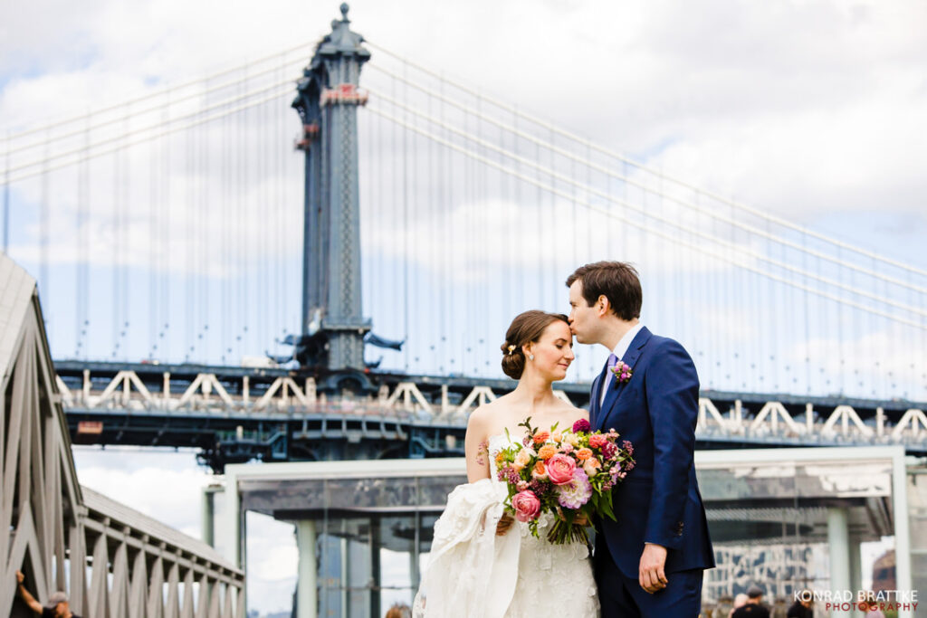 A bride and groom standing in front of the Brooklyn Bridge with the River Cafe in the background.