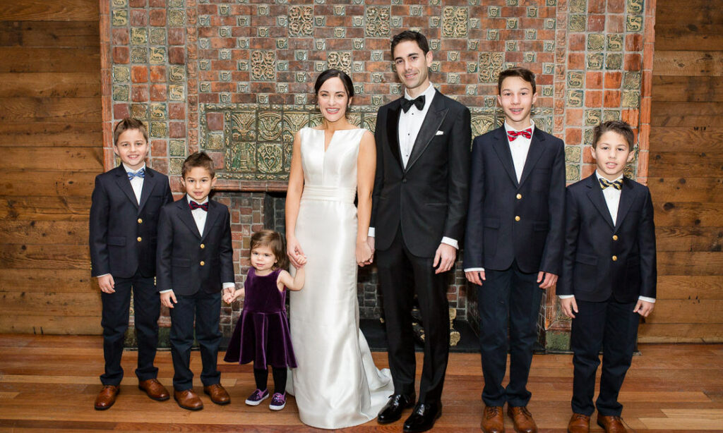 A bride and groom pose with children in front of a fireplace.