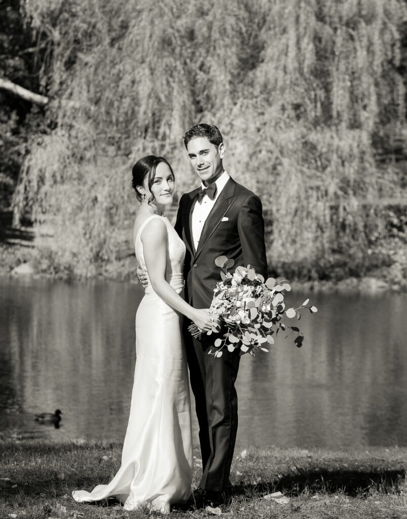 A bride and groom standing in front of a pond in central park.