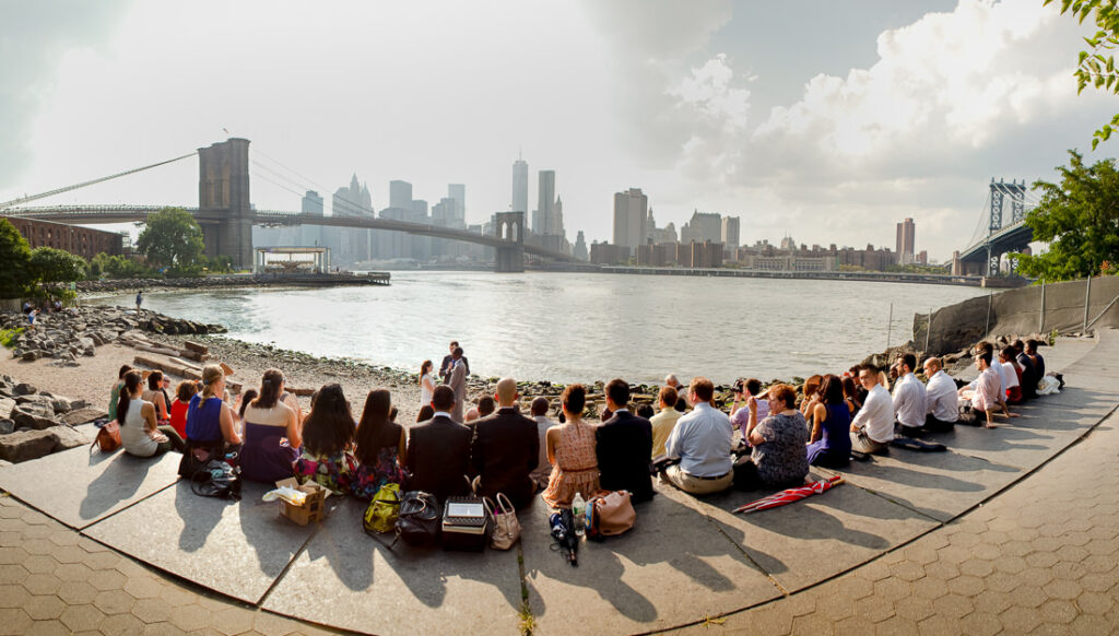 A group of people sitting on a bench in front of the Brooklyn Bridge, enjoying the ceremony.