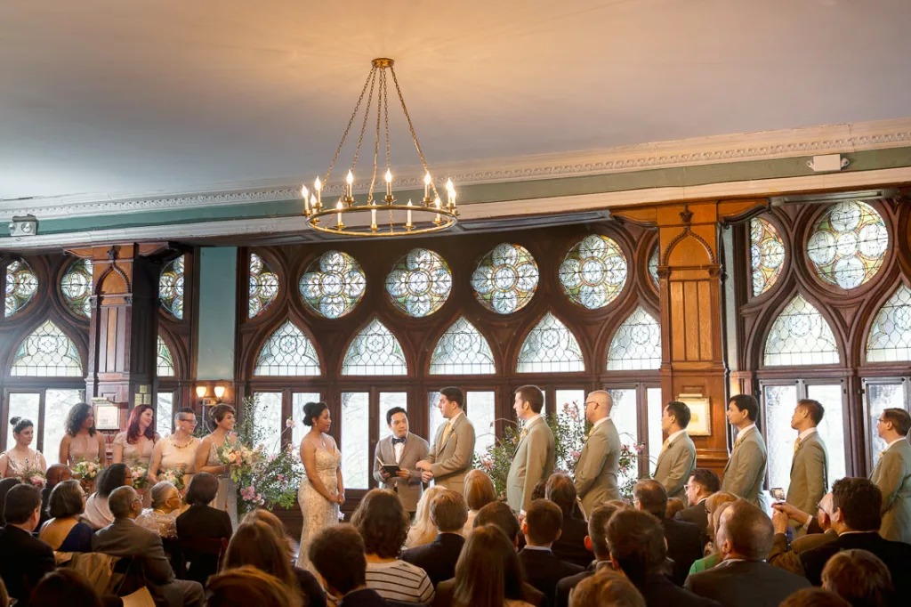 A wedding ceremony in a large room at the Montauk Club with stained glass windows.