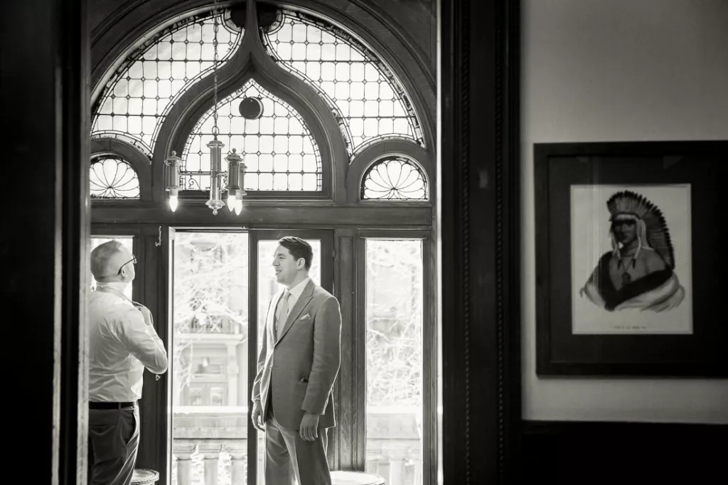 A groom in a suit is standing in front of an ornate door at the Montauk Club.