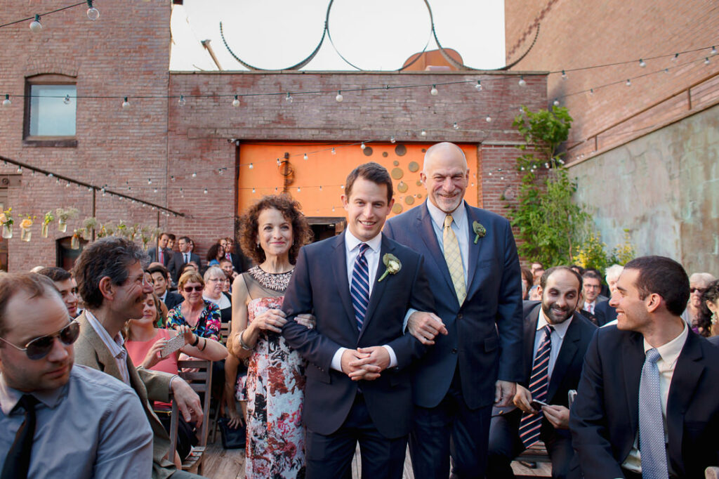 A groom and his parents walking down the aisle under the enchanting light of mymoon at an outdoor wedding.