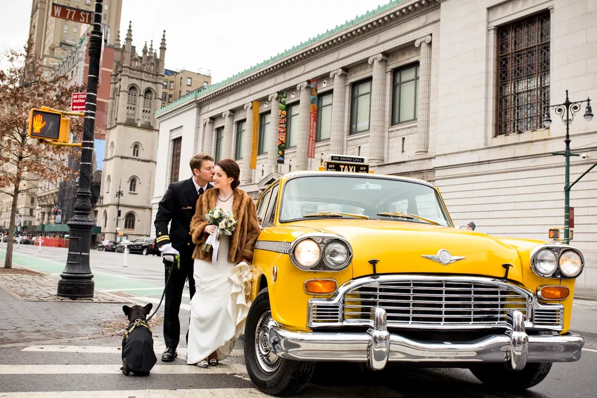 A bride and groom pose next to a yellow taxi.