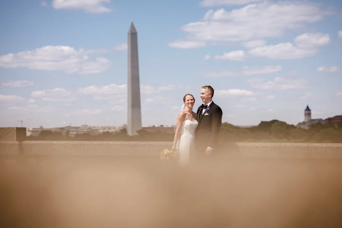 A bride and groom pose outdoors with the Washington Monument in the background under a partly cloudy sky.
