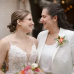 Two brides smiling at each other; one is wearing a white dress and the other a white suit. Both are holding a flower bouquet.