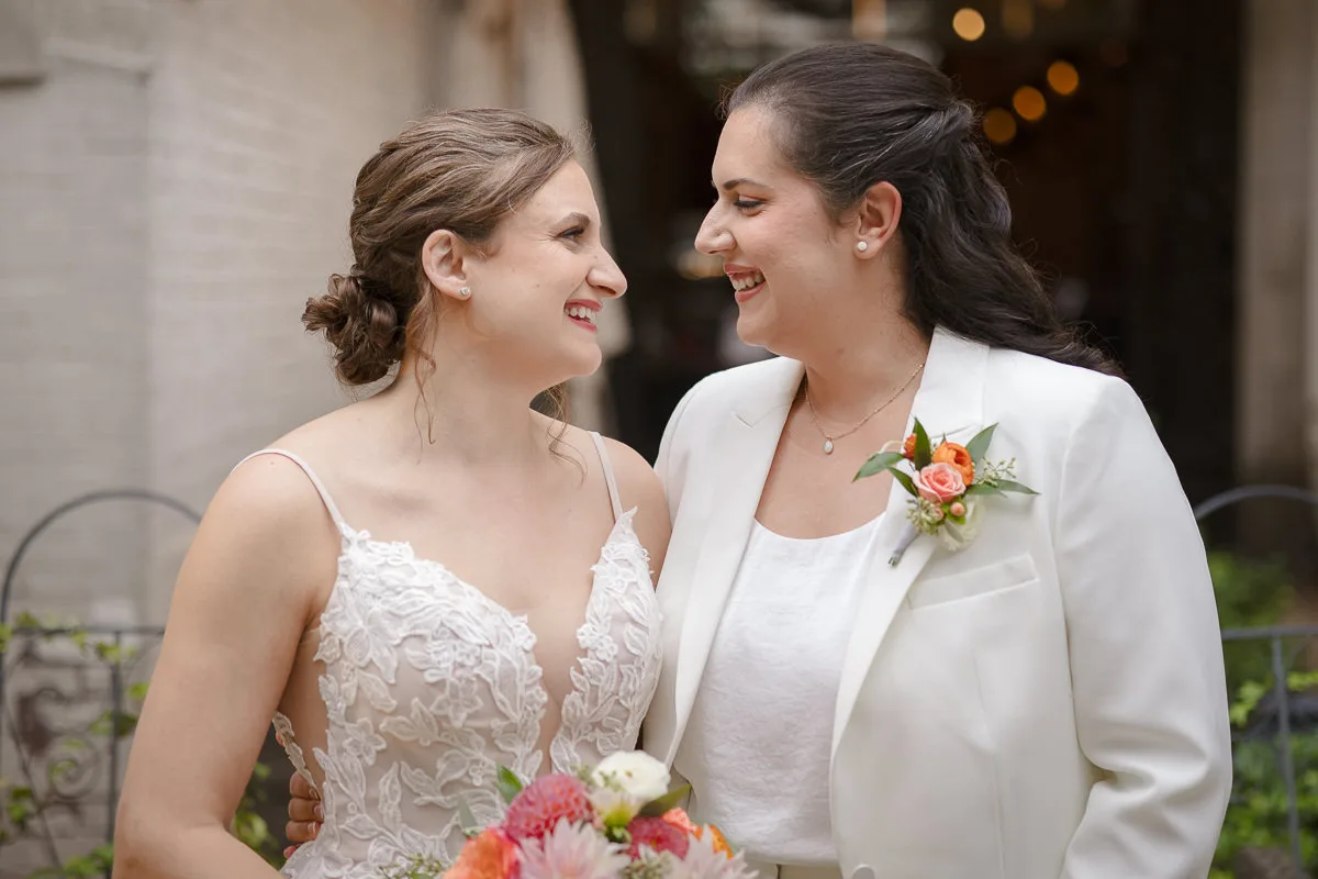 Two brides smiling at each other; one is wearing a white dress and the other a white suit. Both are holding a flower bouquet.