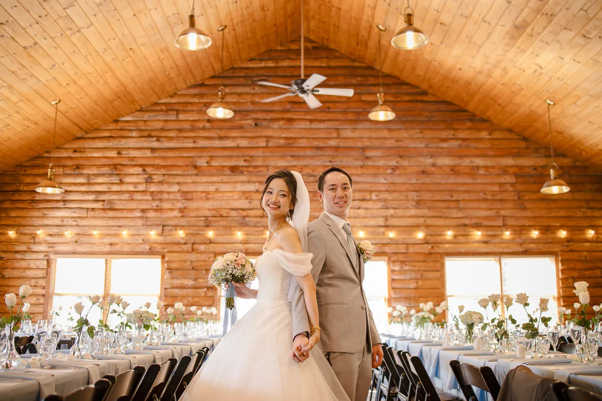 Bride and groom smiling and holding hands in a wooden hall, with decorated tables and hanging lights.