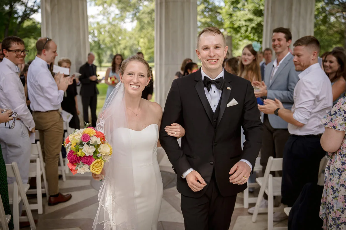 A bride and groom smile while walking down an aisle outdoors. The bride holds a colorful bouquet. Guests are clapping and standing on either side of the aisle.