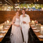 Two brides stand side by side at a long dining table with place settings, flowers, and candles. They are dressed in formal wedding attire, one in a suit and the other in a dress, smiling at the camera.