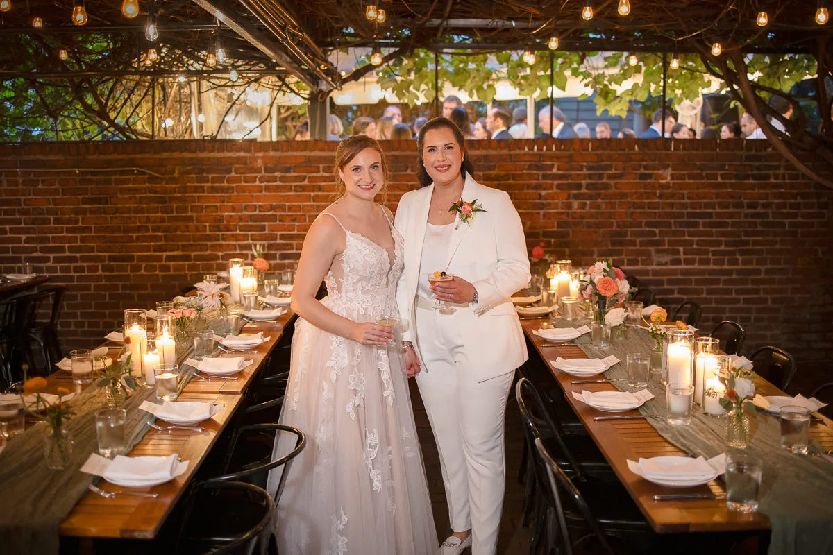Two brides stand side by side at a long dining table with place settings, flowers, and candles. They are dressed in formal wedding attire, one in a suit and the other in a dress, smiling at the camera.